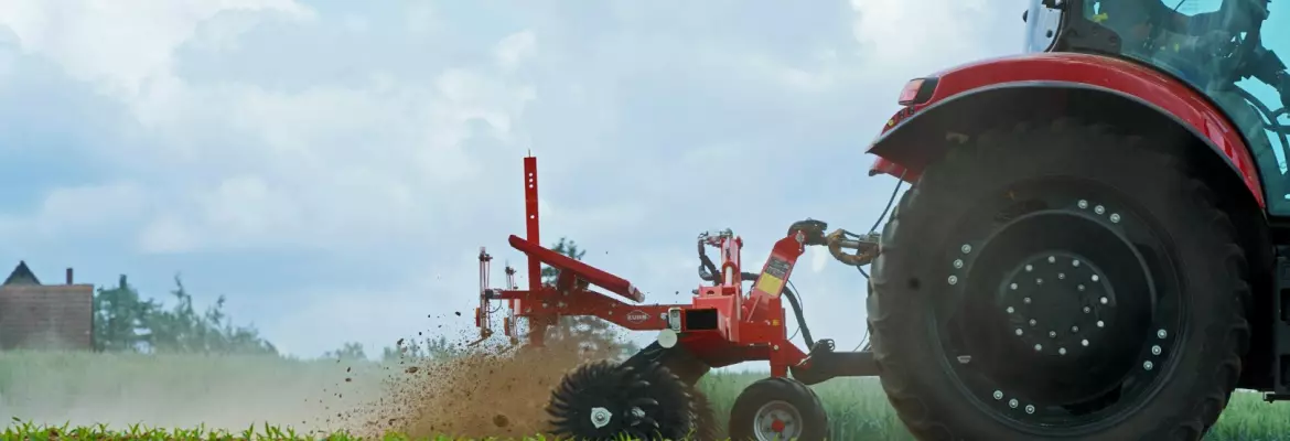 A red tractor pulls Rotary hoe Starliner across a dirt field, kicking up dust. Green crops grow in rows nearby under a cloudy sky with farm buildings visible in the background.
