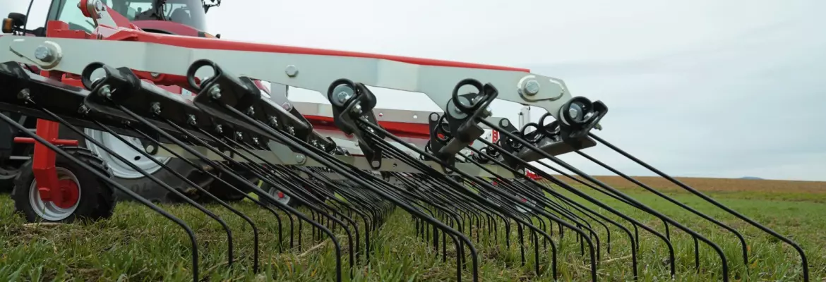 A red and white tractor with a tine weeder Tineliner is working in a grassy field under an overcast sky, preparing soil for farming.