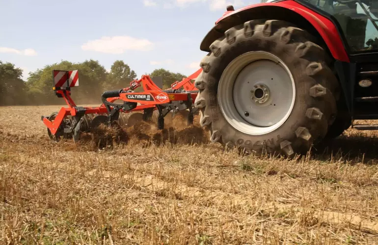 A green tractor pulls a Kuhn stubble tine cultivator Cultimer tillage implement across a harvested field, with dust clouds rising from the soil preparation work under partly cloudy skies.
