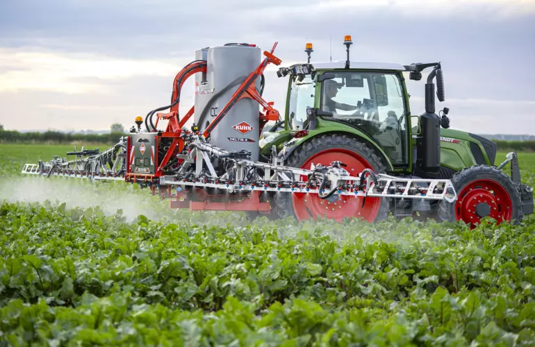 A green tractor with DELTIS mounted sprayer operates a large agricultural spraying implement across a field of green crops, creating visible spray mist under a cloudy sky.