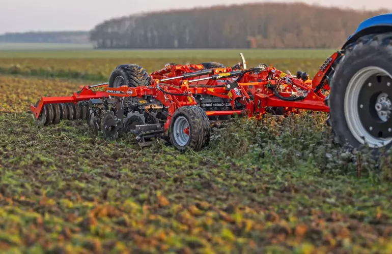 Kuhn's stubble disc cultivator Optimer XL 5000 being pulled by a tractor across a ploughed field, with rolling hills and forests visible in the background under an overcast sky.