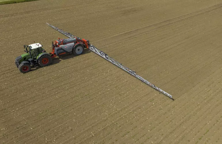 A green tractor pulling KARAN an efficient trailed sprayer across a brown agricultural field with visible furrow lines.