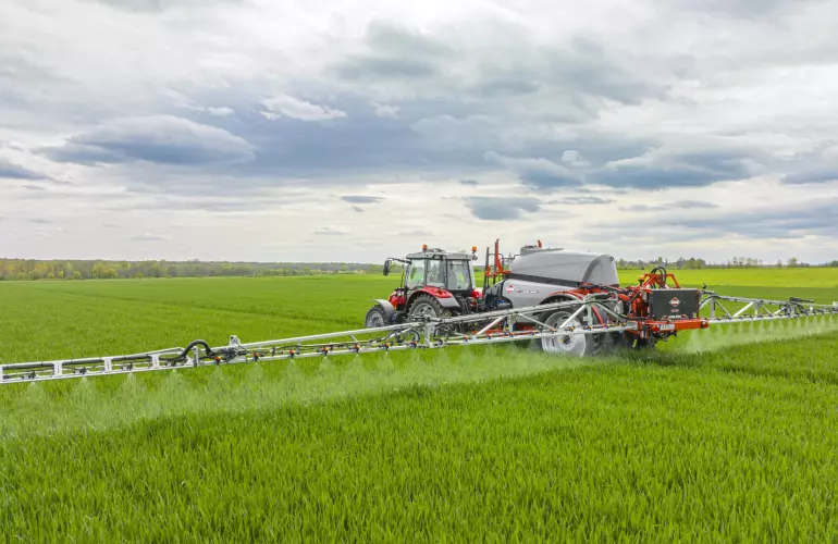 A red tractor with trailed sprayer LEXIS during sprayin operation on a vibrant green field under a cloudy sky, with trees visible on the horizon.