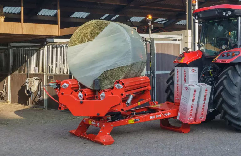 A KUHN SW1114 wrapping a round bale in front of a horse stable.