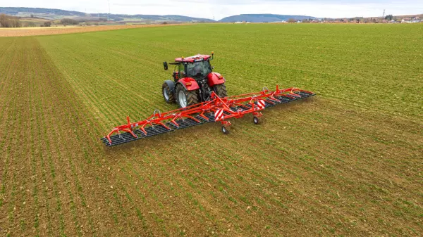 A red tractor pulls Kuhn's tine weeder Tineliner across a green field with visible crop rows, surrounded by rolling hills and distant farmland under an overcast sky.