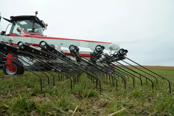 A red and white tractor with a tine weeder Tineliner is working in a grassy field under an overcast sky, preparing soil for farming.