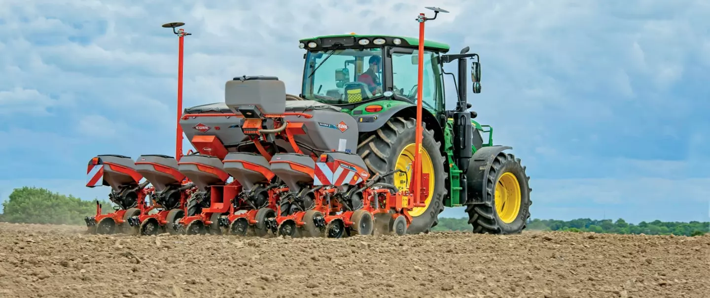 A green tractor pulls Kuhn's precision seed drill MAXIMA across a tilled dirt field under a cloudy blue sky with forested areas in the background.
