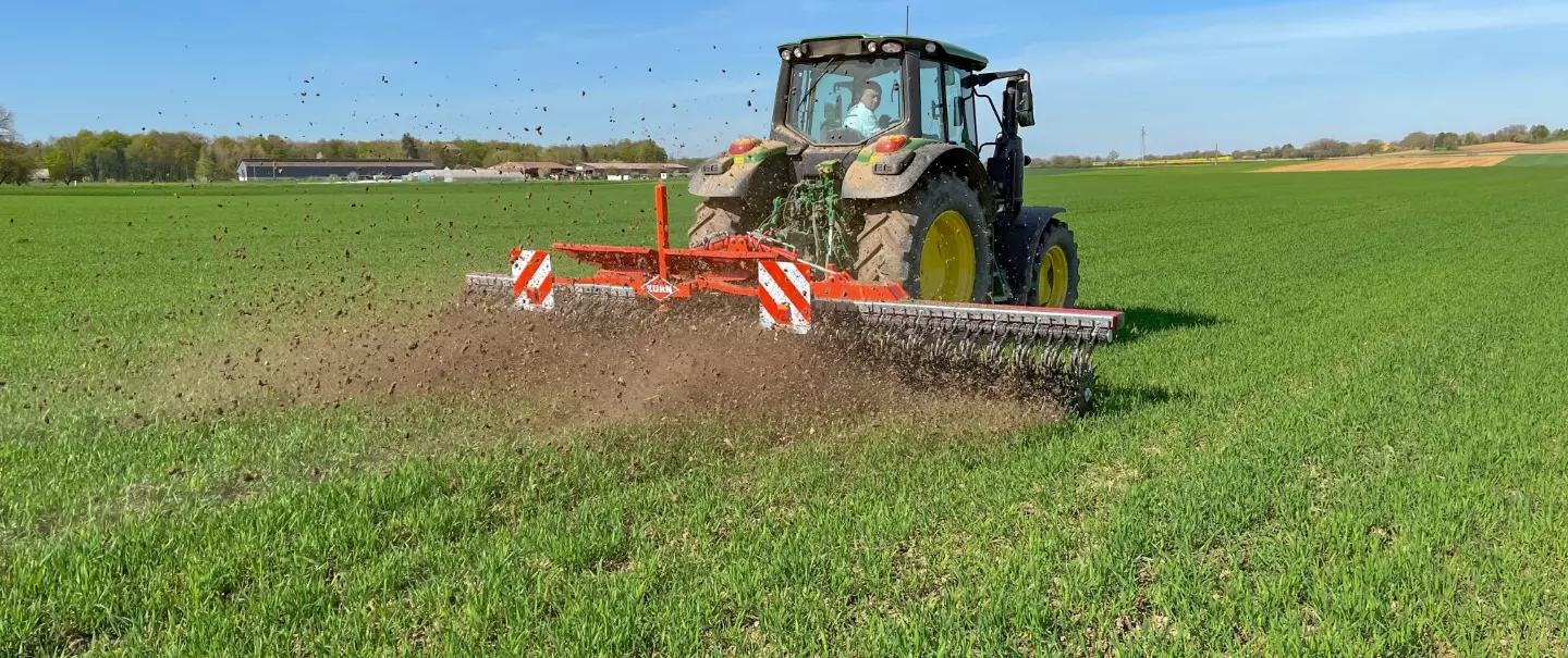 A tractor pulls red Kuhn rotary hoe Starliner across a green field, tilling soil in strips. The scene shows agricultural work under a clear blue sky with birds visible overhead.