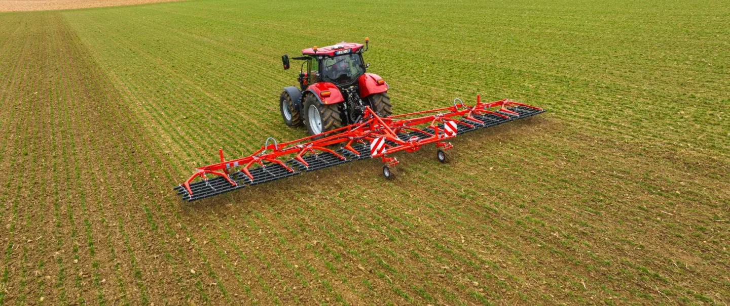 A red tractor pulls Kuhn's tine weeder Tineliner across a green field with visible crop rows, surrounded by rolling hills and distant farmland under an overcast sky.